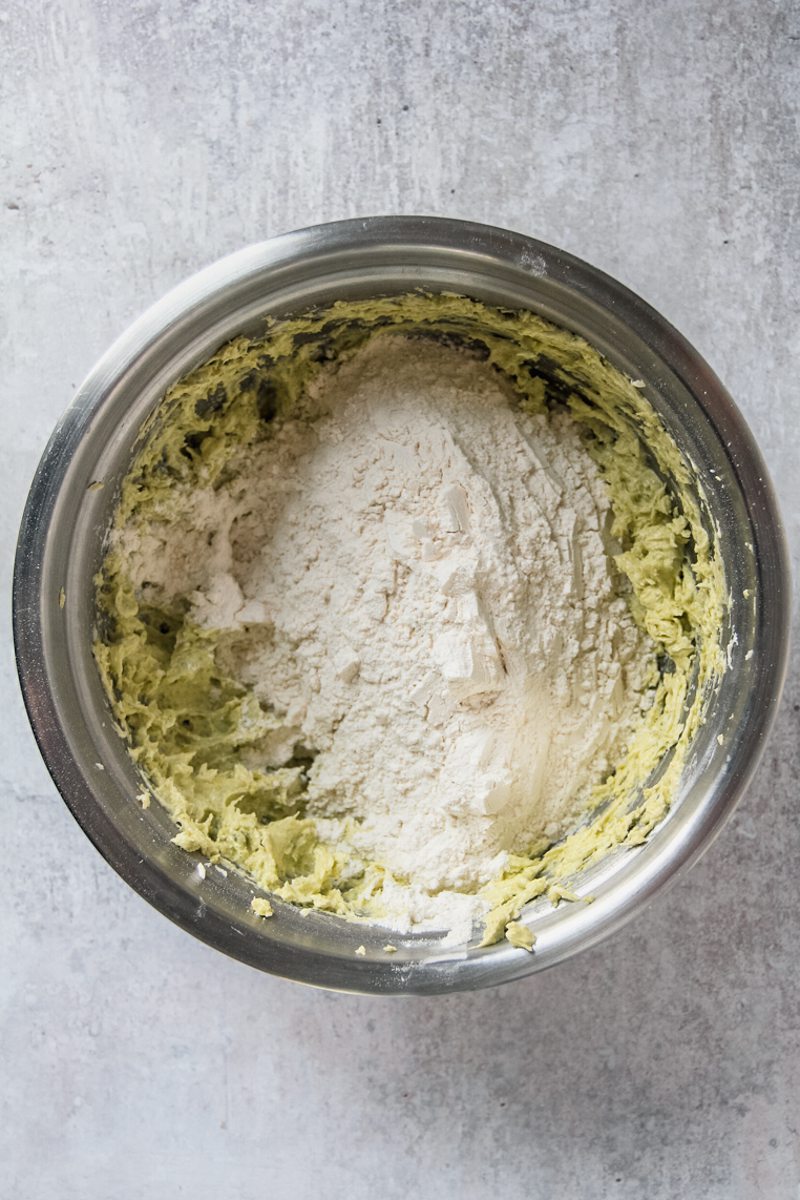 Dry ingredients sit on top of a pistachio sugar cookie dough mixture in a stainless steel bowl on a gray surface.