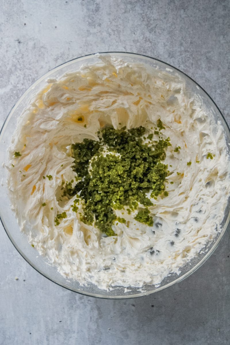 Pistachio paste sits on top of a buttercream mixture in a glass bowl on a gray surface.