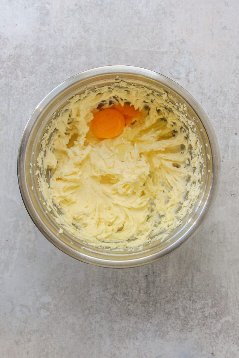 An egg sits on top of a whipped cookie base in a stainless steel bowl on a gray surface.