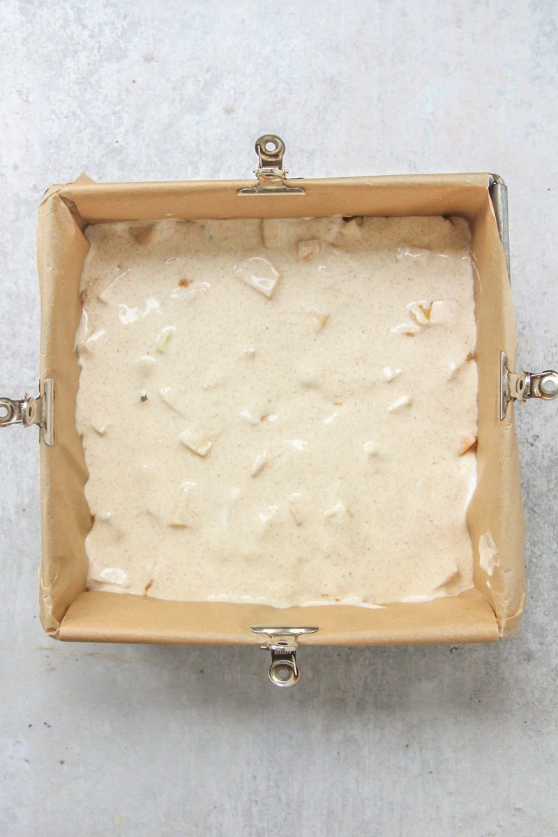 A loose cheesecake batter mixture sits on top of apples in a lined square tin on a gray surface.