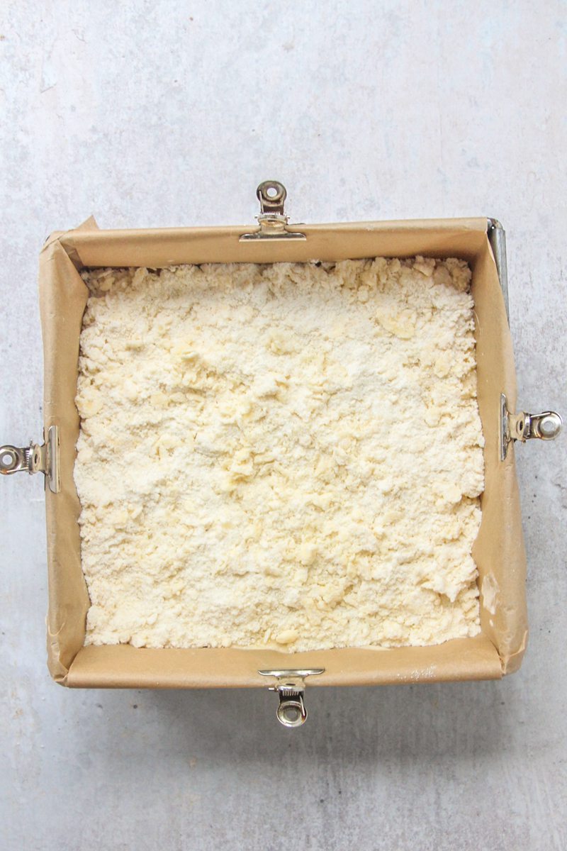 A streusel mixture sits on top of an apple cheesecake mixture on a square lined tin on a gray surface.