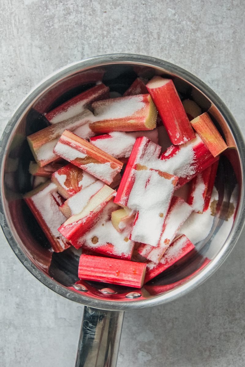 Cut stalks of rhubarb sit in a stainless steel saucepan on a gray surface coated in sugar.