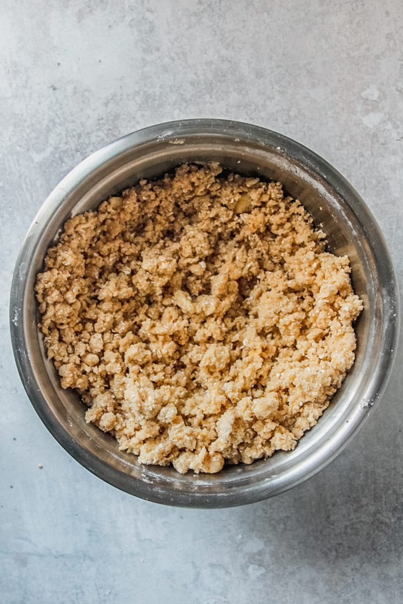 A rough oat and flour crumble mixture sits in a stainless steel bowl on a gray surface.