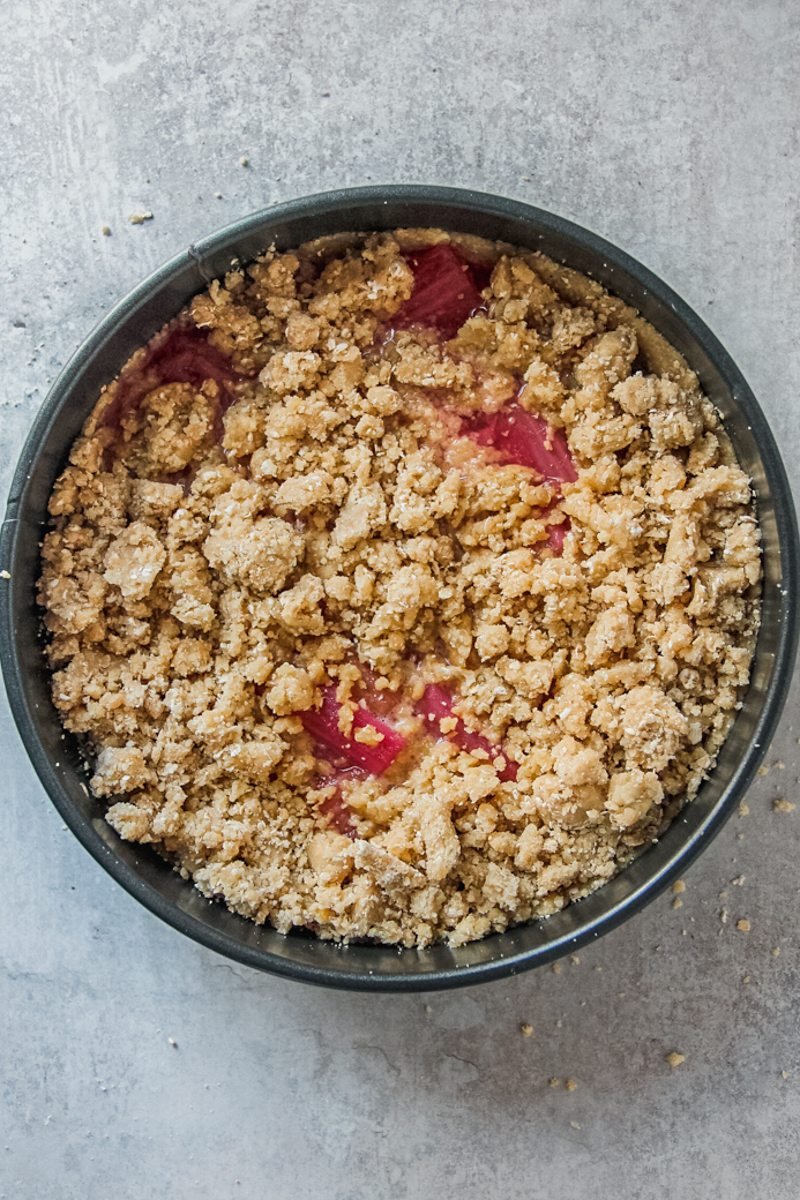 A crumble mixture sits on top of rhubarb pieces in the center of a cake in a metal cake tin on a gray surface.