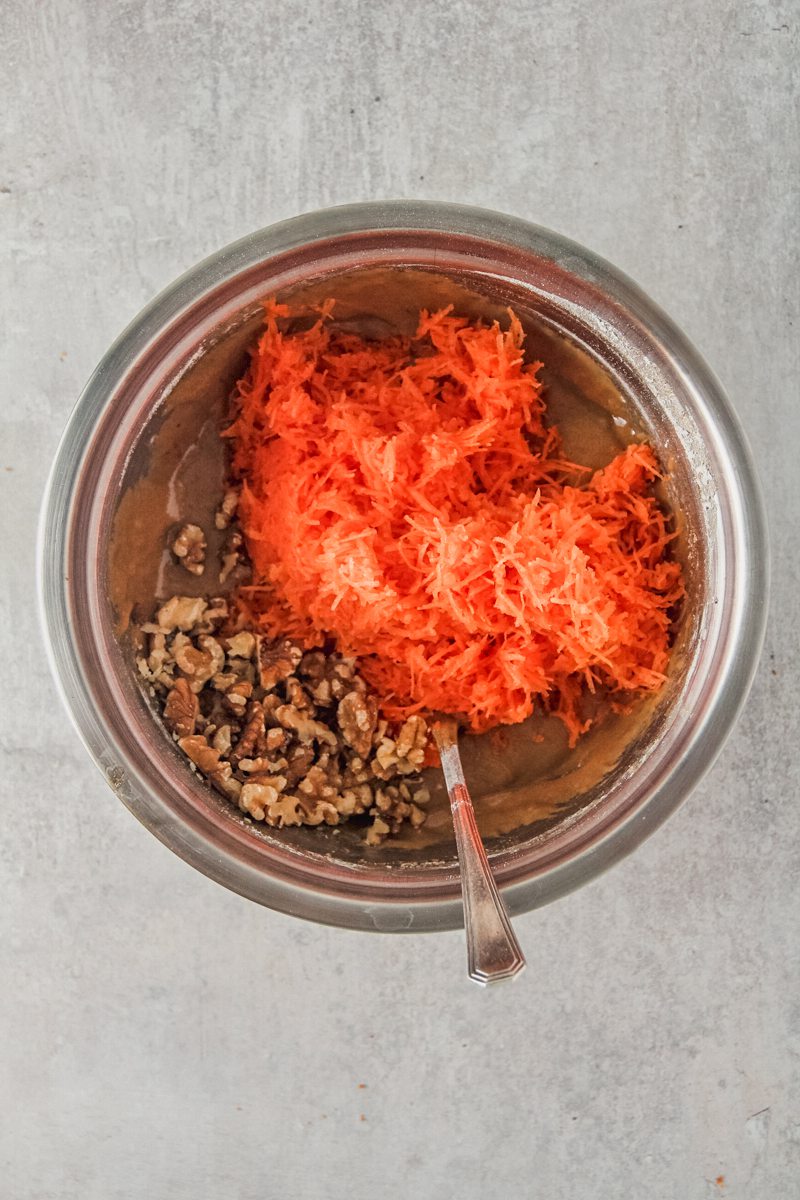 Grated carrot and walnuts sit on top of carrot cake batter in a stainless steel bowl on a gray surface.