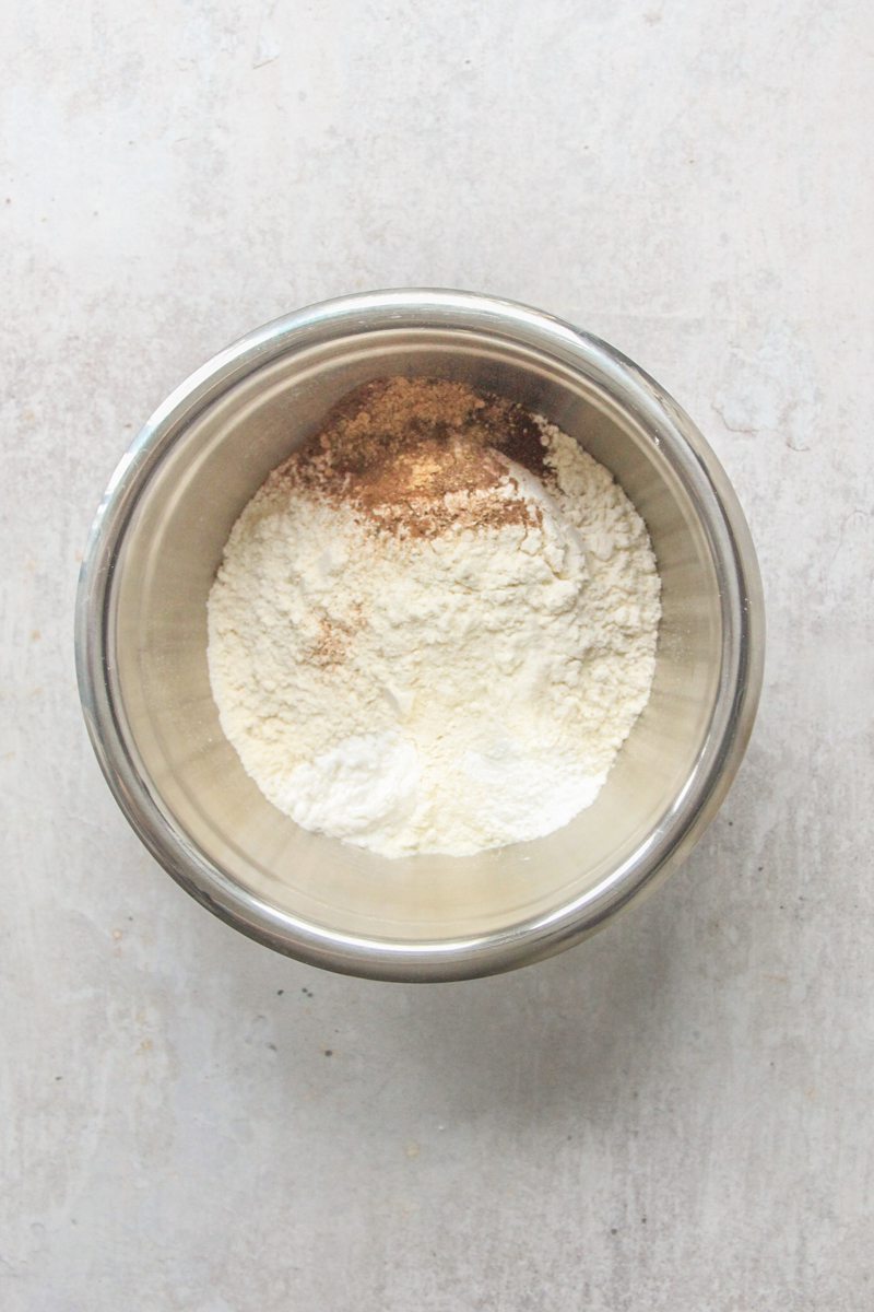 Dry ingredients, including ground spices sit in a stainless steel bowl on a gray surface.
