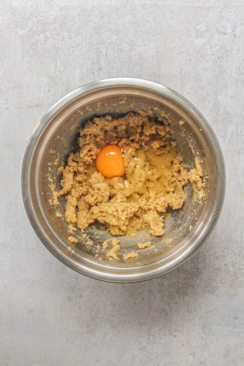 An egg sits on a whipped butter and sugar mixture in a stainless steel bowl on a gray surface.