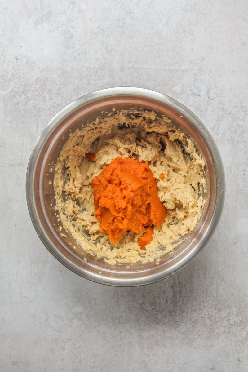 Pumpkin puree sits on top of a whipped butter, sugar and egg mixture in a stainless steel bowl on a gray surface.