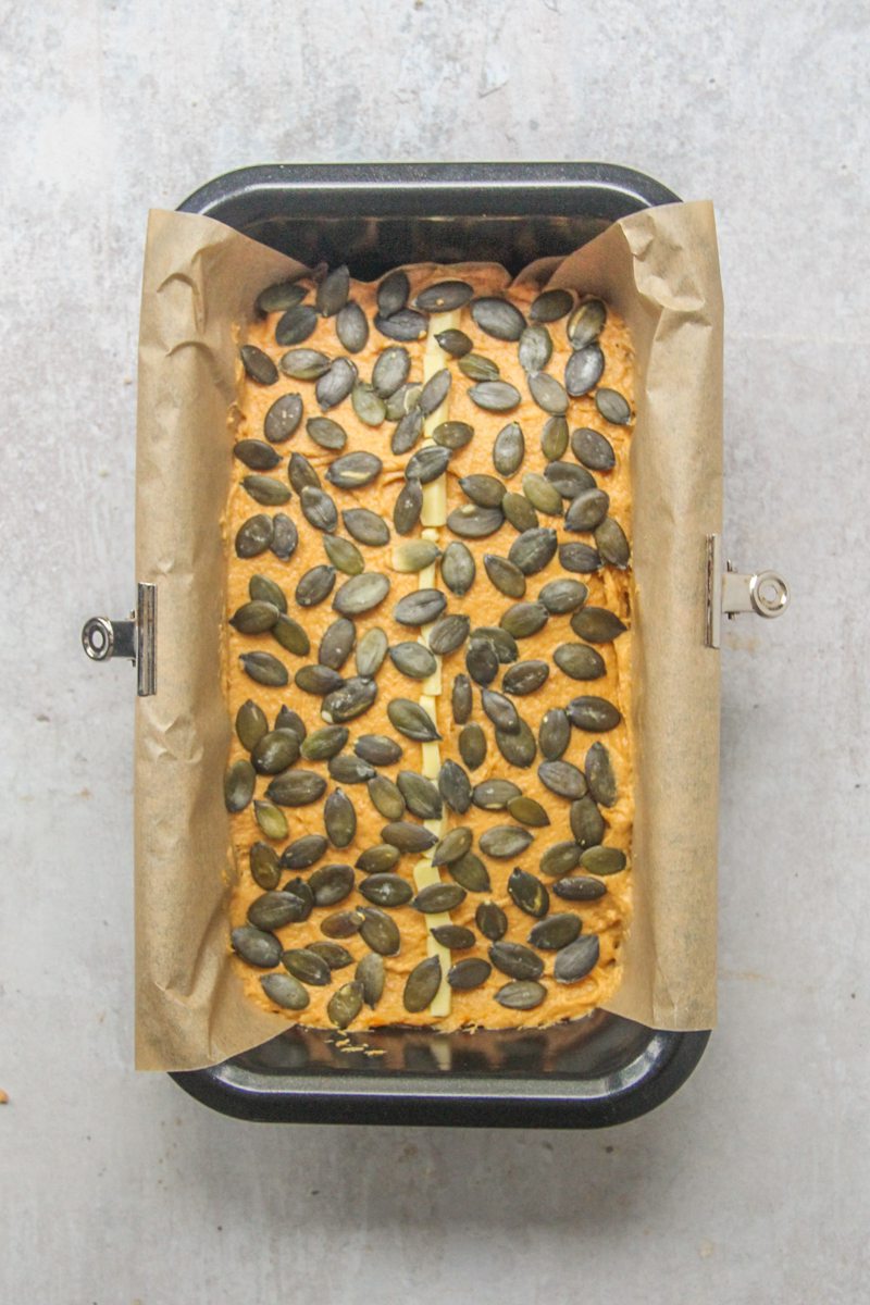 Pumpkin seeds sit on top of unbaked pumpkin bread batter in a black metal loaf tin on a gray surface.
