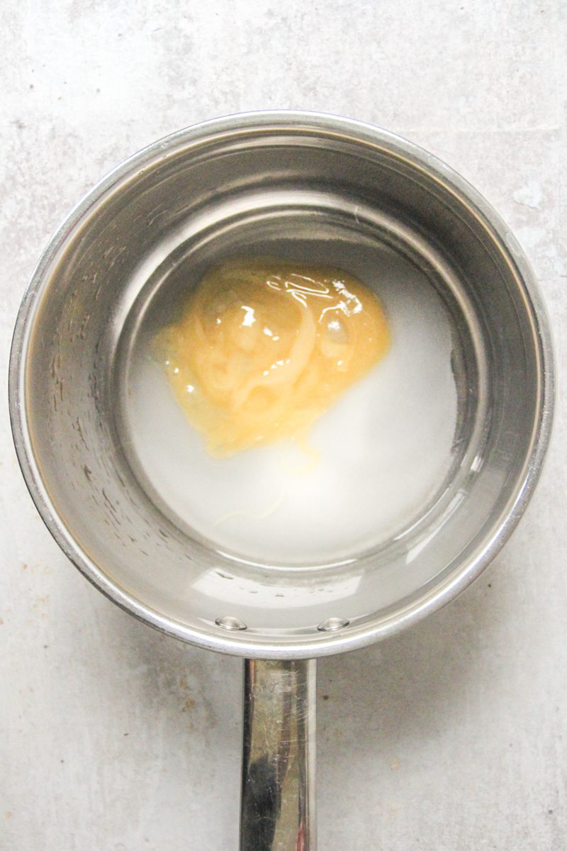 Honey, sugar and water sit in a stainless steel saucepan on a gray surface.