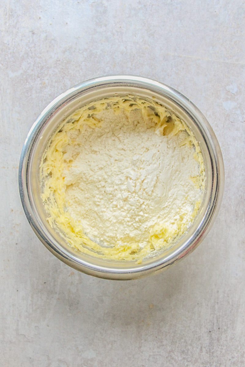 Dry ingredients sit on top of whipped cookie dough in a stainless steel bowl on a gray surface.
