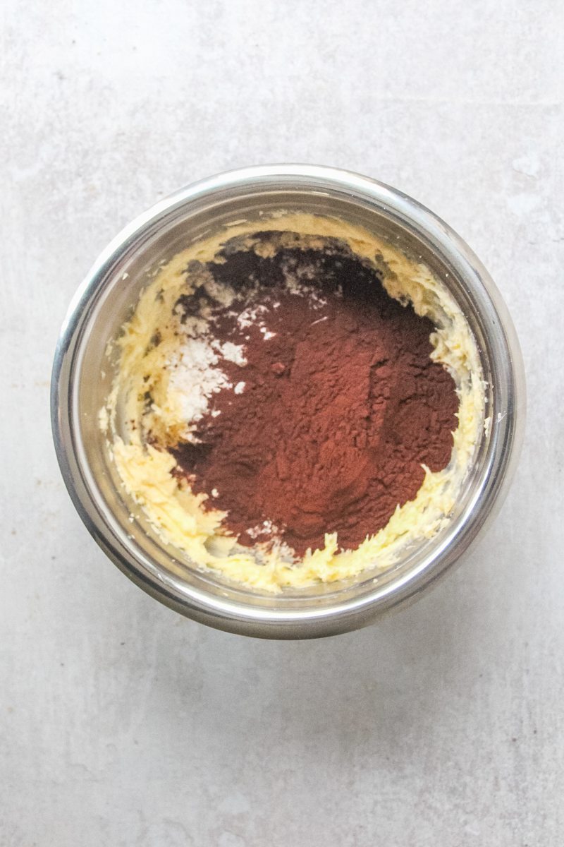 Flour and cocoa powder sits on top of a whipped butter mixture in a stainless steel bowl on a gray surface.