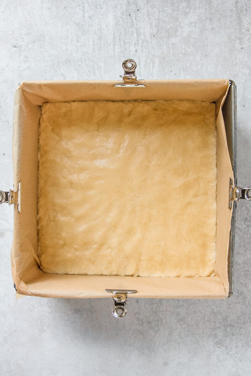 A sugar cookie bar mixture sits pressed into a lined square metal baking tin on a gray surface pre baking.