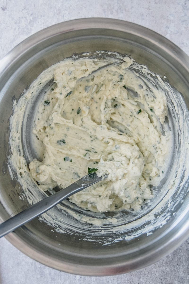 A creamy cheese mixture with herbs sits stirred in a stainless steel bowl on a gray surface.