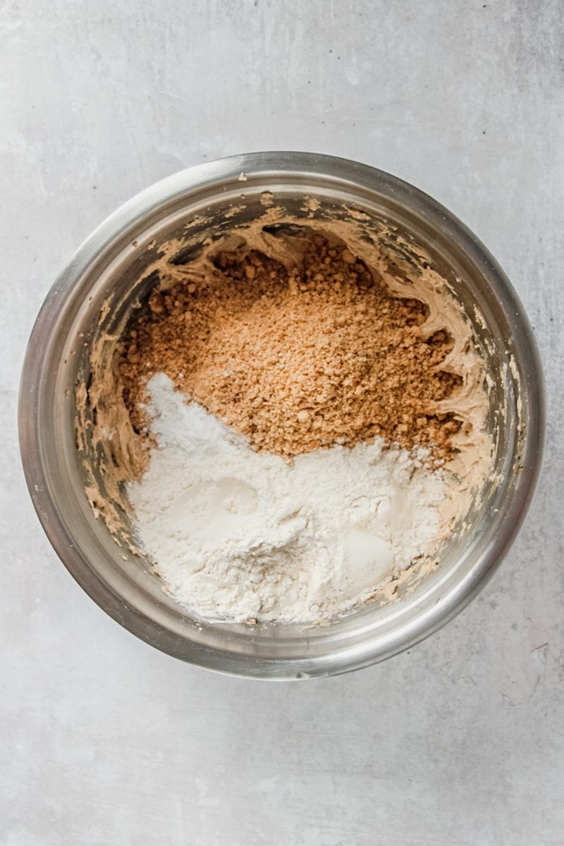 Crushed graham crackers or digestvies and flour sit on top of cookie dough in a stainless steel bowl on a gray surface.