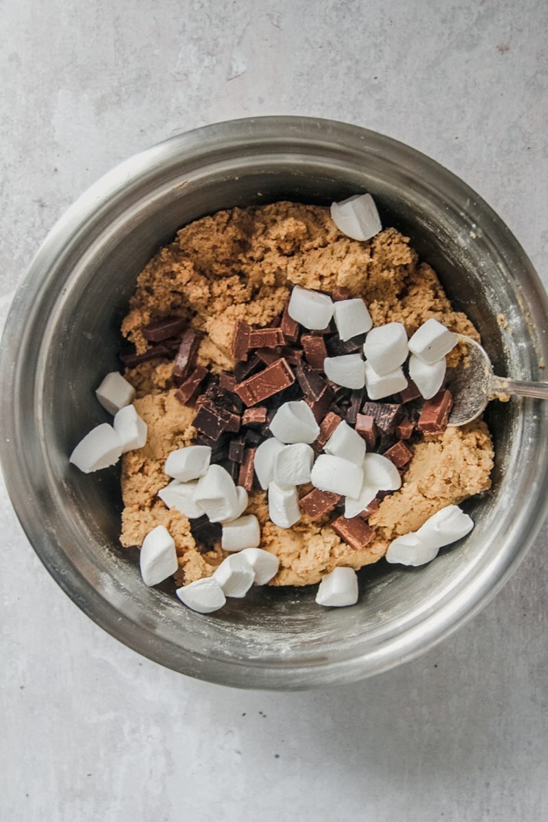 Marshmallows and chocolate chunks sit on top of cookie dough in a stainless steel saucepan on a gray surface.