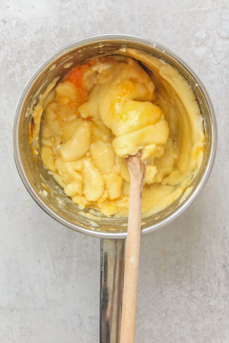 Eggs sit in a flour, butter and water mixture in a stainless steel saucepan on a gray surface.