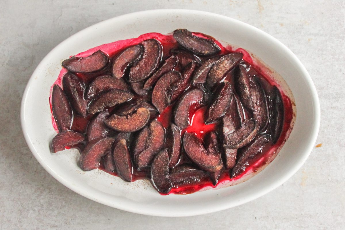 Roasted plums in a white oval baking dish on a gray surface.