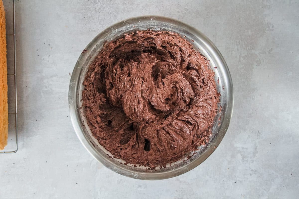 Chocolate buttercream sits in a stainless steel bowl before being placed on top of a coconut cake on a light grey background.