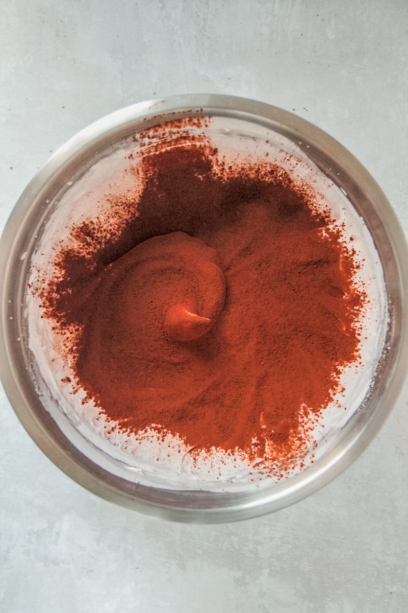 Cocoa powder sits on top of whipped meringue in a stainless steel bowl on a gray surface.