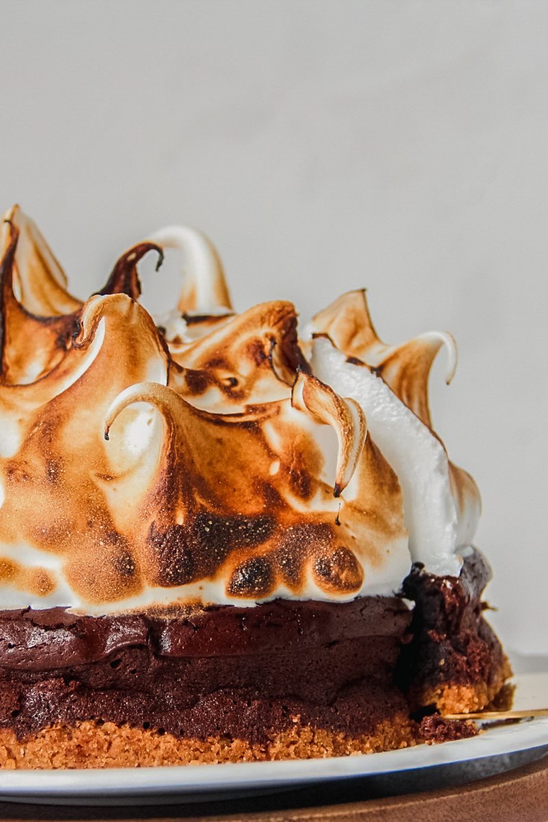 A close up of smore pie sitting on a white ceramic plate on a wooden surface with a sliver of the marshmallow interior visible from the visible piece of pie sitting on a cake server.