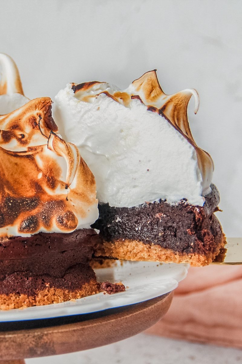 A close up of smore pie sitting on a white ceramic plate on a wooden surface with the interior visible from the visible piece of pie sitting on a cake server.