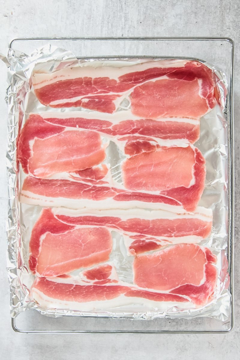 Strips of bacon sit on aluminum foil on a baking tray on a gray surface.