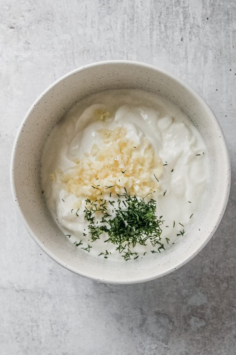 Yogurt sits in a bowl with finely chopped herbs and garlic on a gray surface.