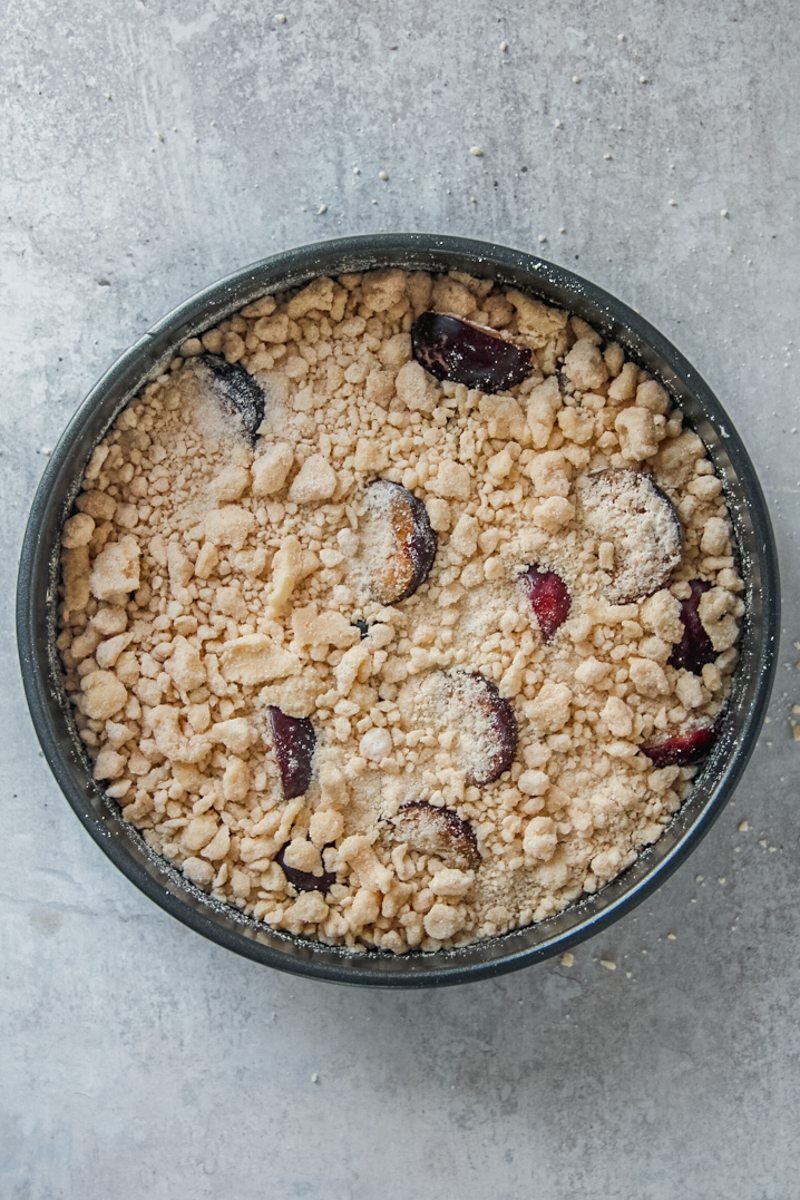 A cake topped with streusel sits inside in a round metal tin on a gray surface.