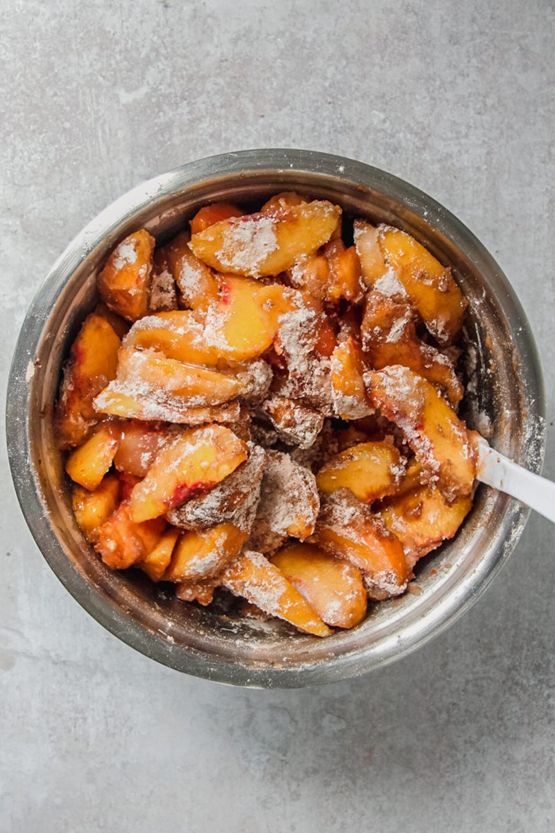 Peaches are stirred with other cobbler filling ingredients for a double crusted pie in a stainless steel bowl on a gray surface.