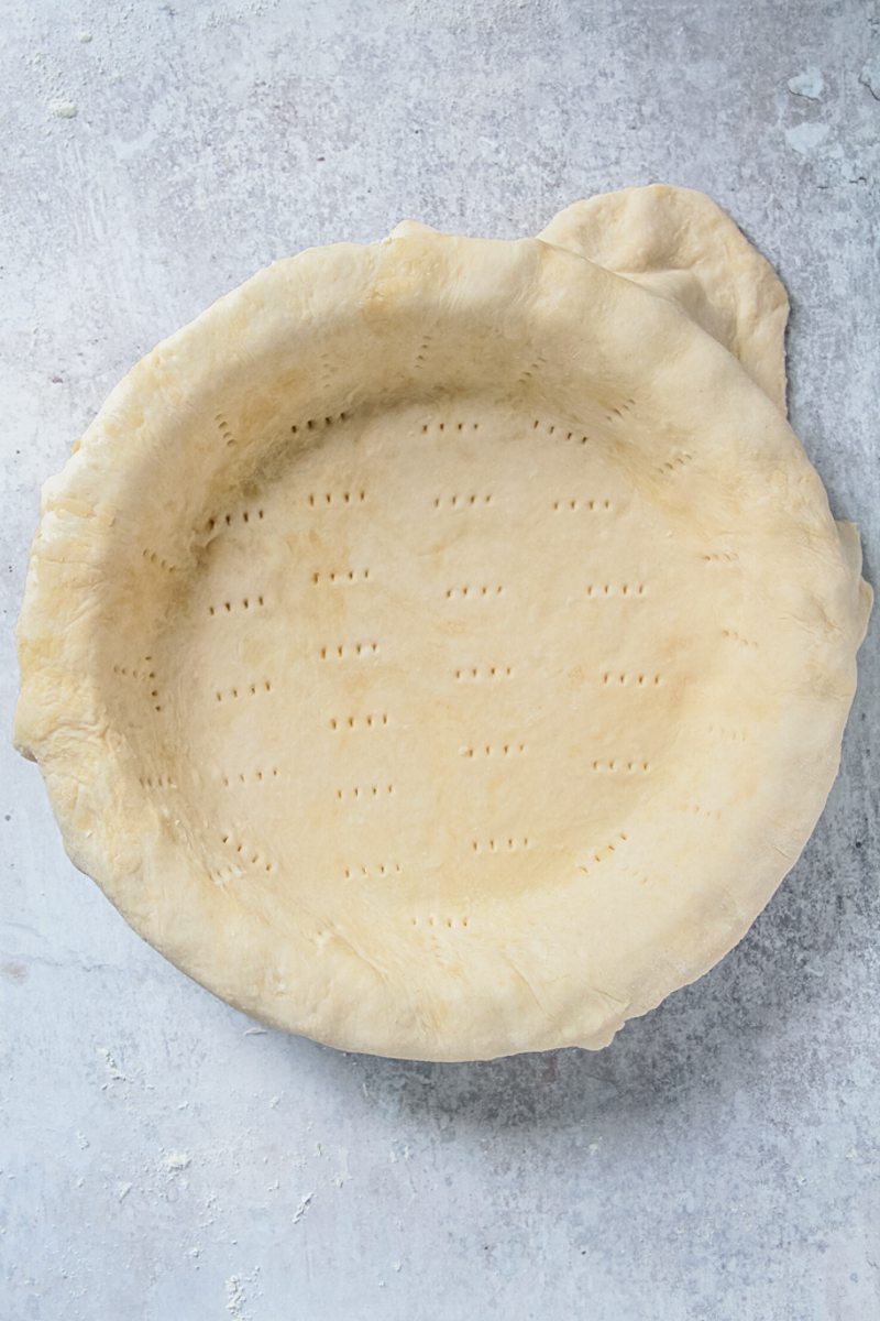 Unbake pie pastry sits in a pie dish with fork prongs pressed therein on a gray surface.