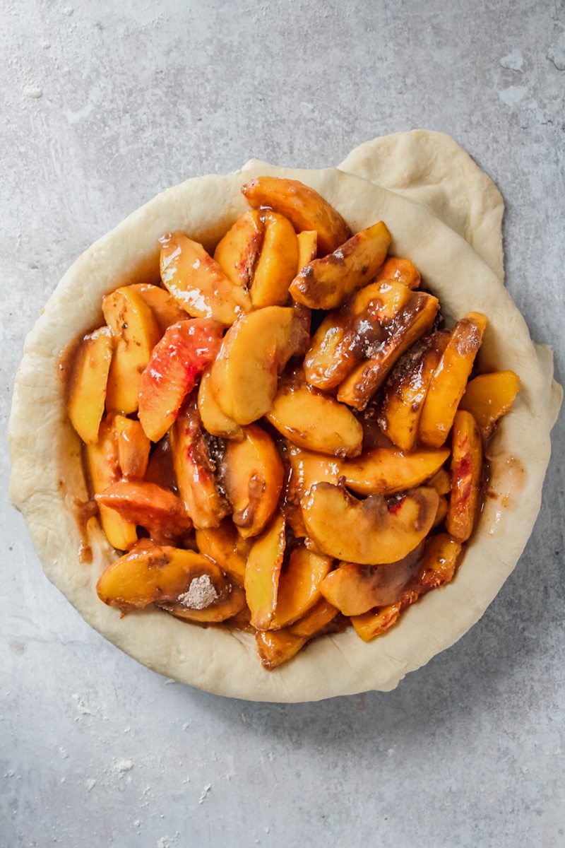 Slices of peaches covered in sugar and cornstarch sit in the base of a pie pastry on a gray surface.