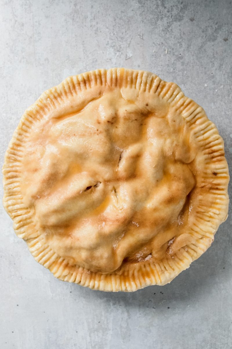 An unbaked double crusted peach cobbler with a fork prong edge and egg glaze topped with sugar and ground cinnamon sits on a gray surface.