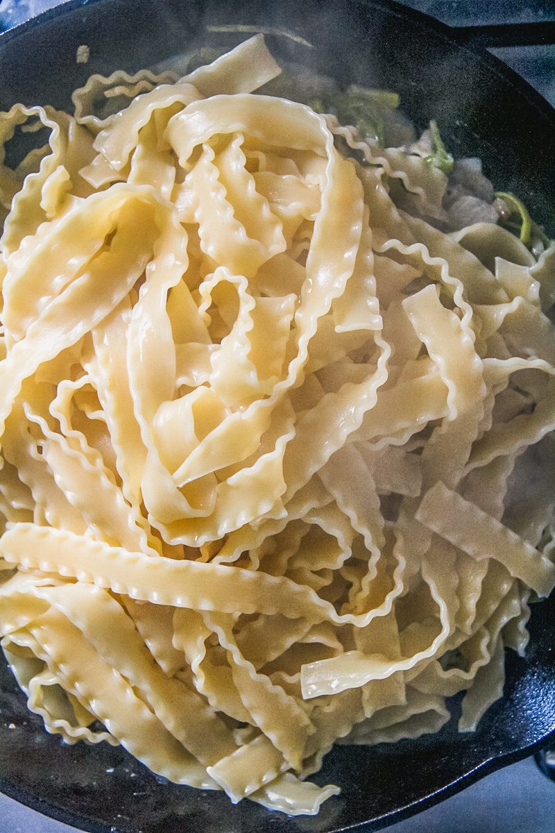 Cooked mafalda pasta sits on top of cooked leeks and goats cheese on a cast iron skillet on a cooker.