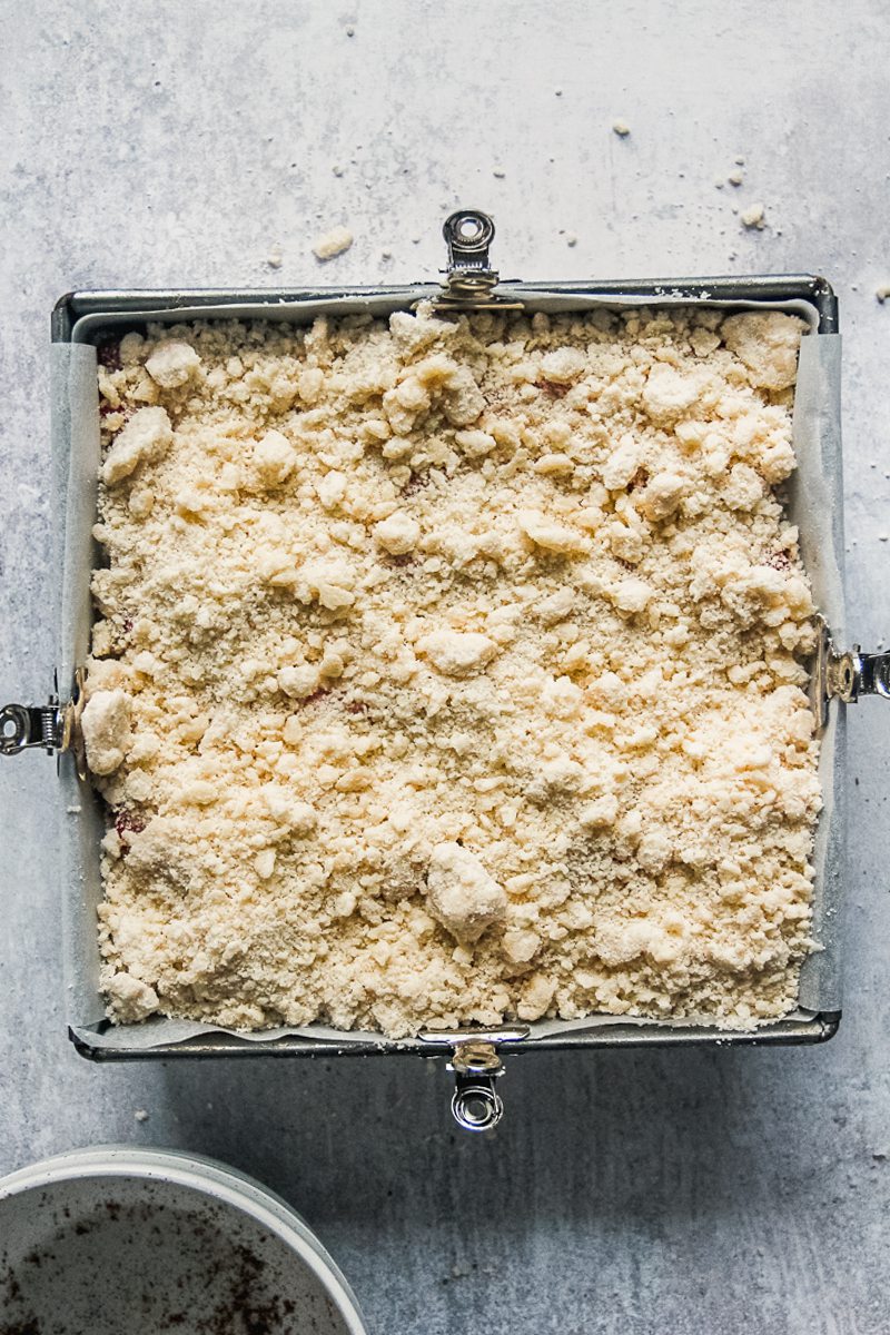 Unbaked streusel sits on top of rhubarb and cheesecake in a lined square metal baking tin.