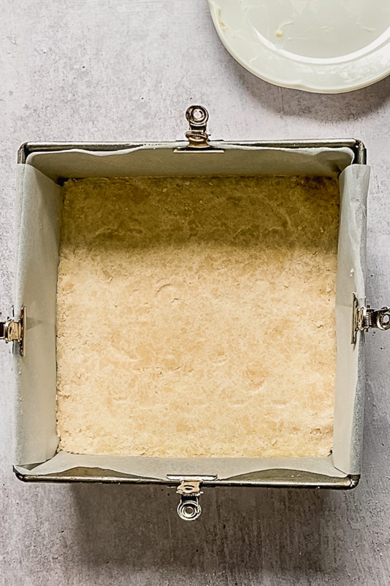 Shortbread crust sits pressed into a lined square metal baking tin on a gray surface.
