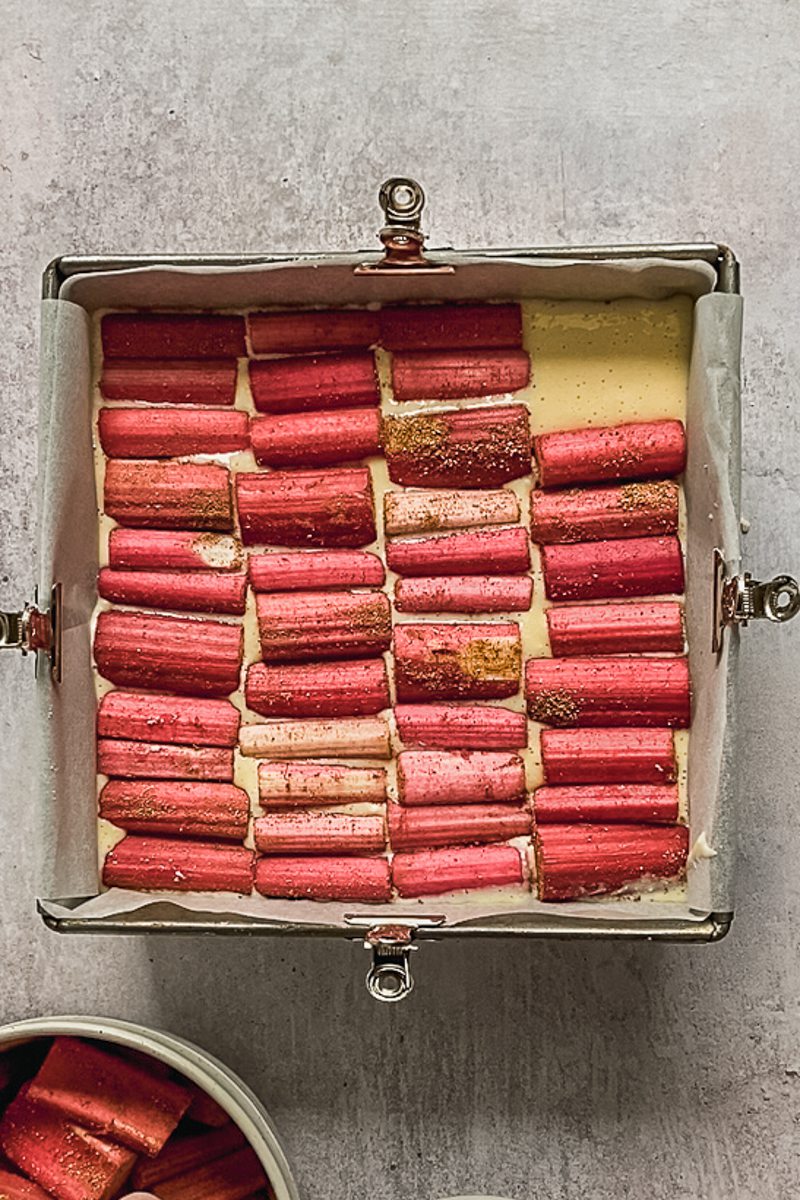 Rhubarb sits on top of cheesecake in a lined square metal baking tin on a gray surface.