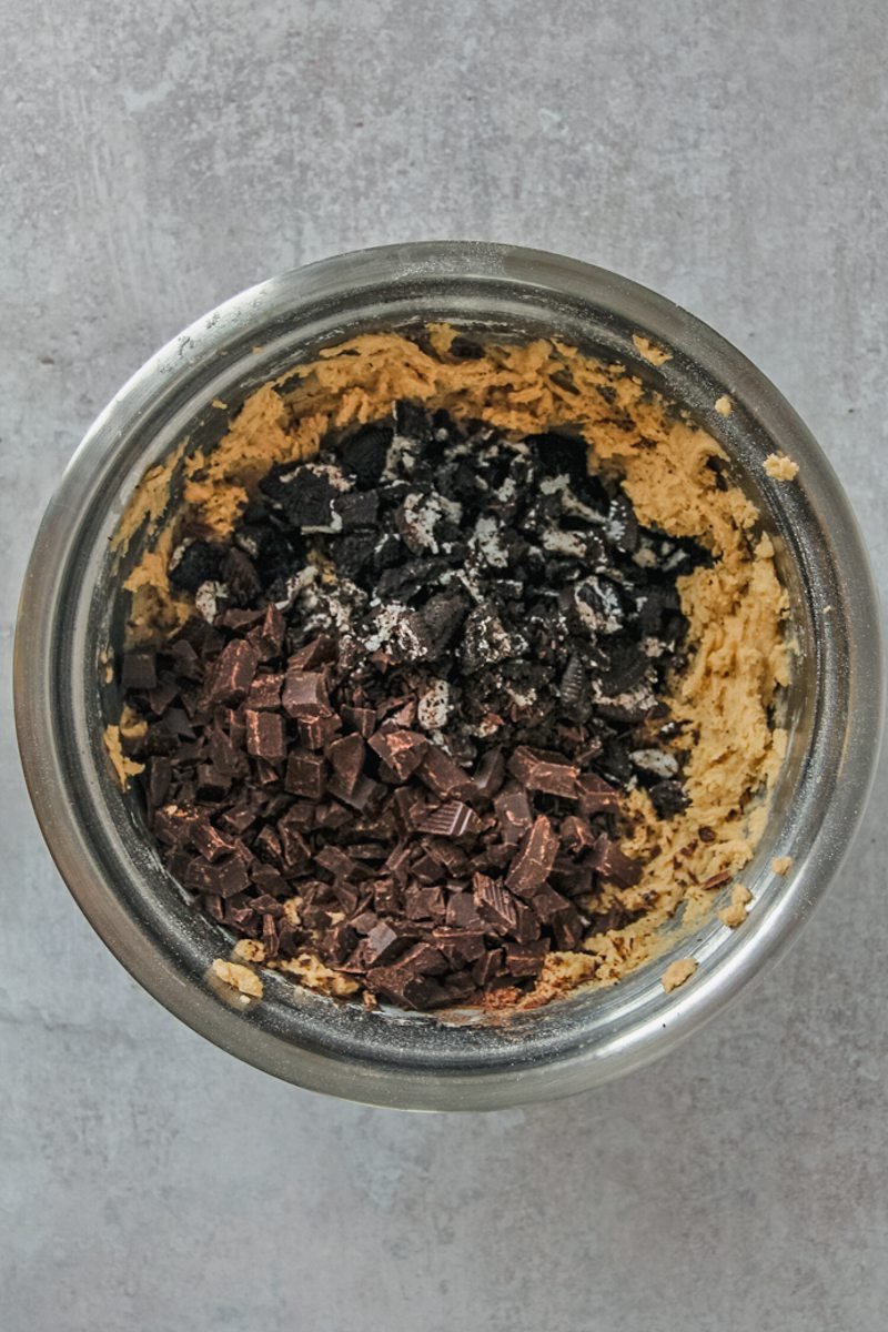 Roughly broken oreo pieces and chocolate chunks sit on top of cookie dough in a stainless steel saucepan on a gray surface.