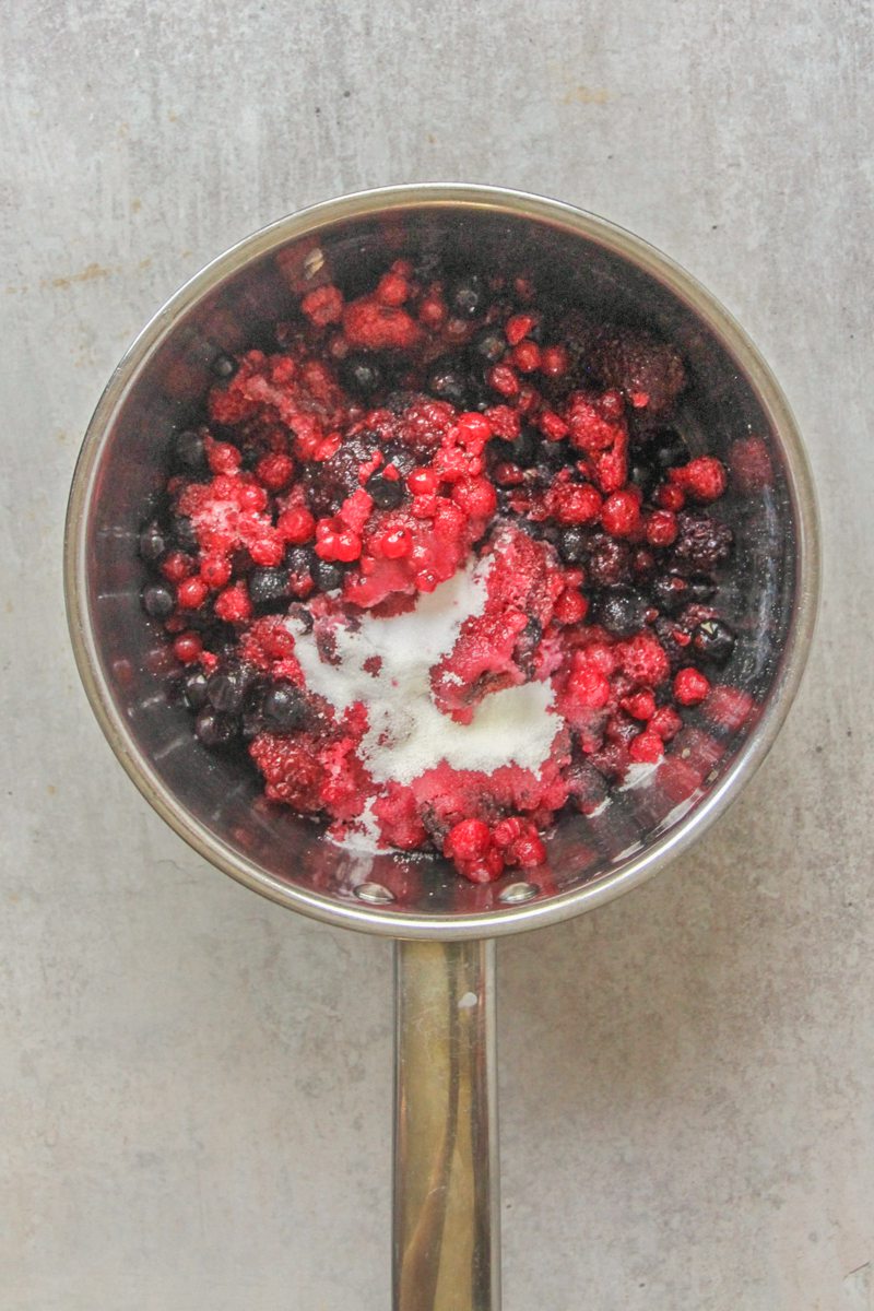 Mixed berries and sugar sit in a stainless steel saucepan on a gray surface.