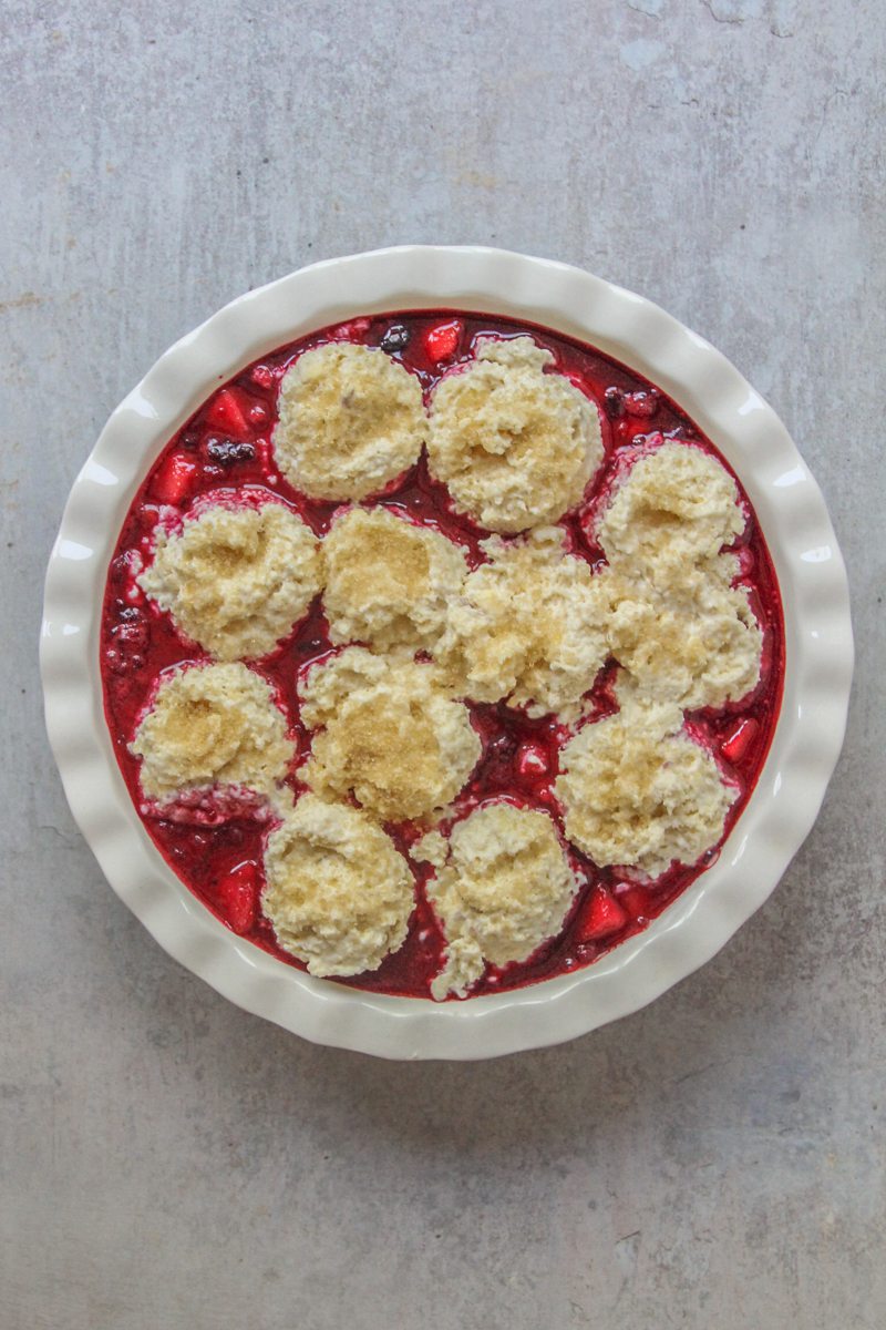 Unbaked apple berry cobbler sits on a white ceramic pie dish on a gray surface.