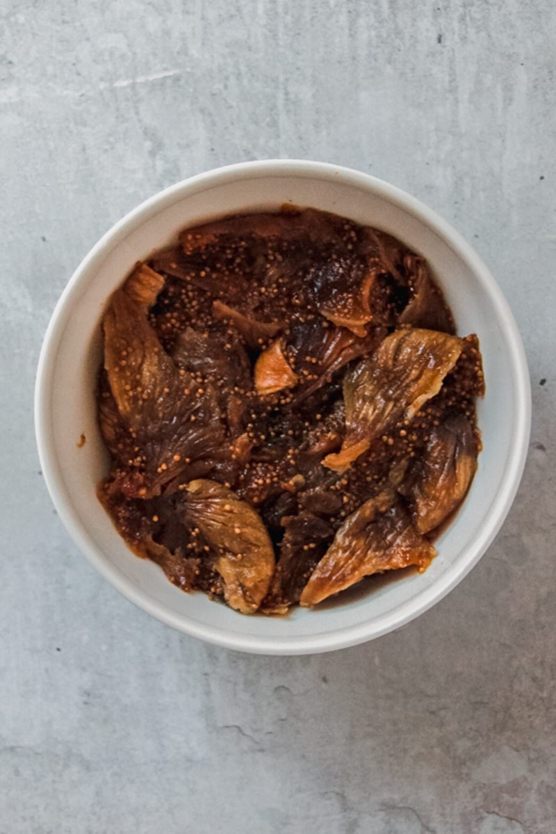 Dried figs soak in boiling water in a white ceramic bowl on a gray surface.