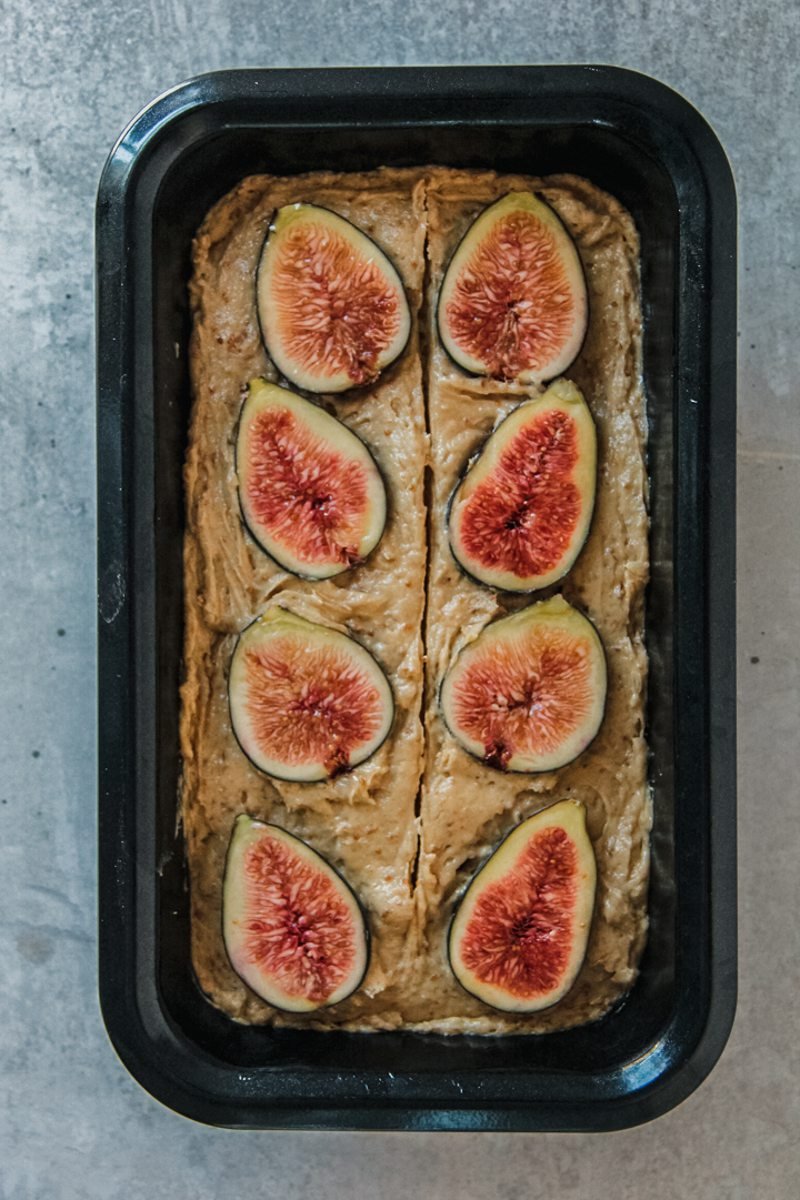 Fig cake sit in a metal loaf tin with fresh figs on top on a gray surface.