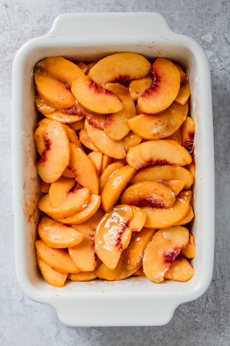 Sliced peaches sit in a white rectangle baking dish stirred with sugar and spices on a gray surface.