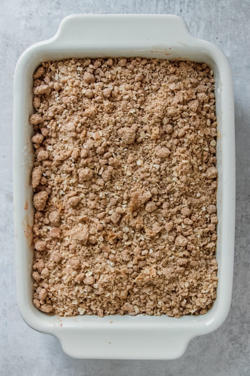 A streusel mixture sits on top of sliced peaches on a white rectangle baking dish on a gray surface.