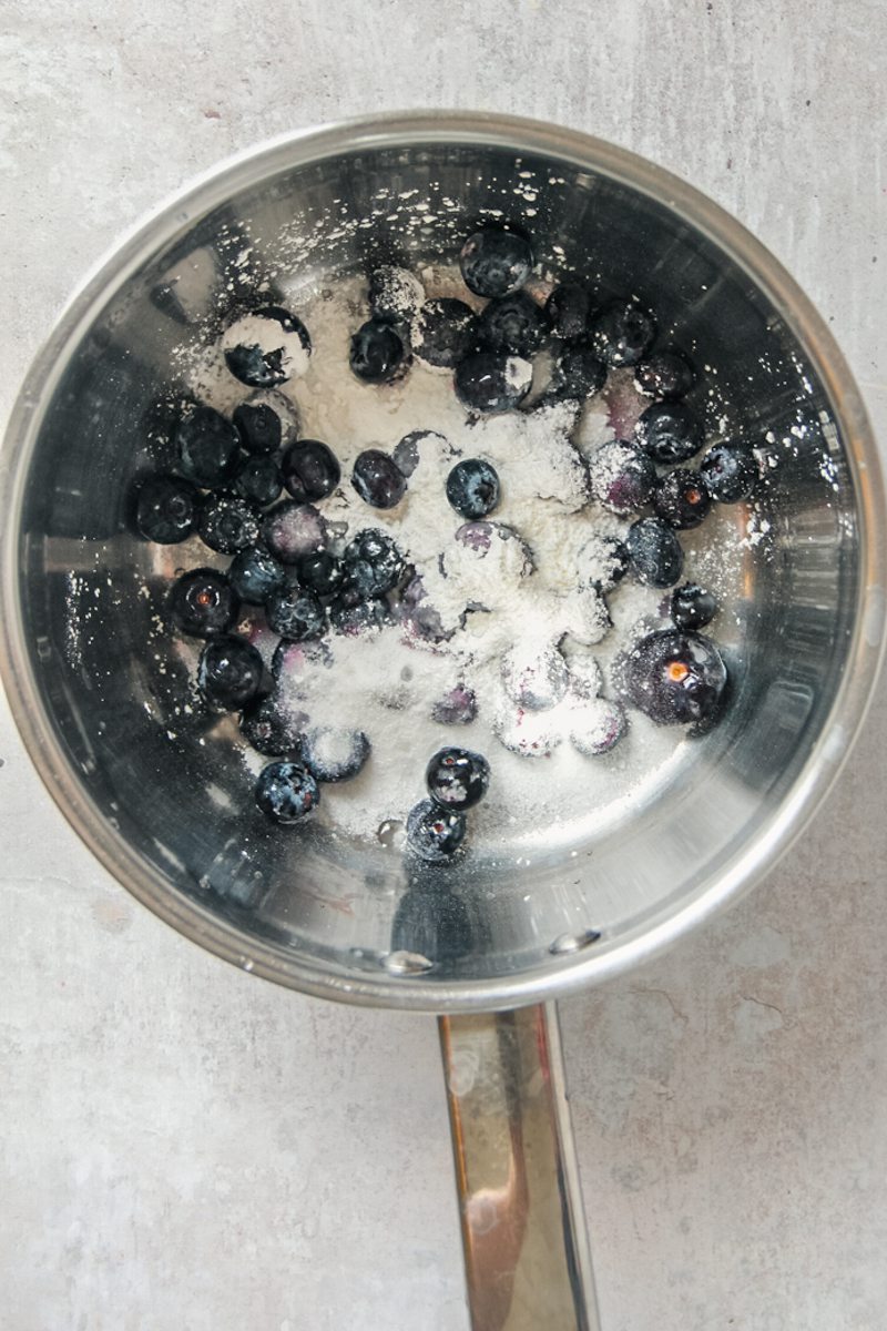 Blueberries, sugar and corn starch sit in a stainless steel saucepan on a gray surface.