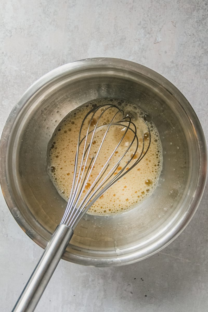 An egg and sugar mixture sits whisked in a stainless steel bowl on a light gray surface.