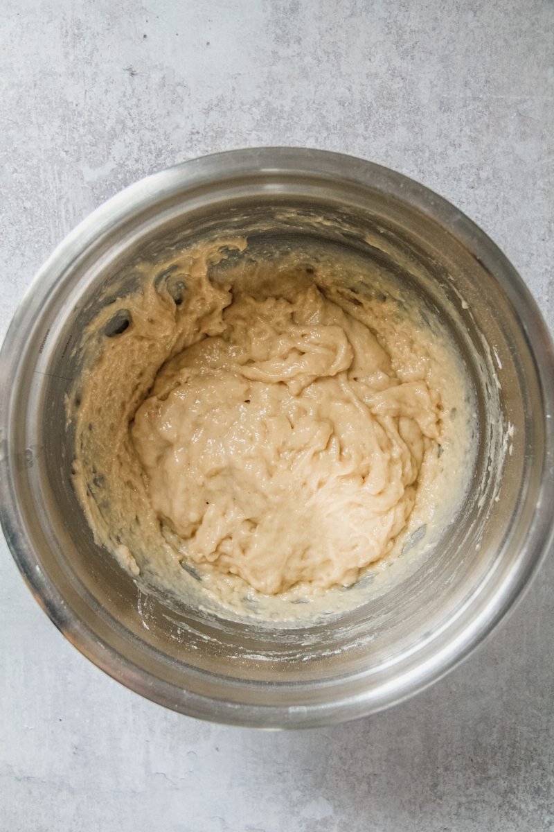 An apple fritter batter sits in a stainless steel bowl on a light gray surface.