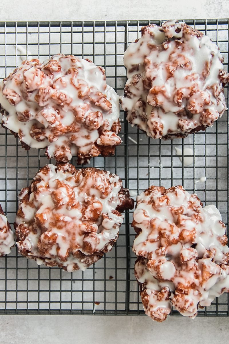 Glazed apple fritters sit on a cooling tray on a gray surface whilst the glaze sets.