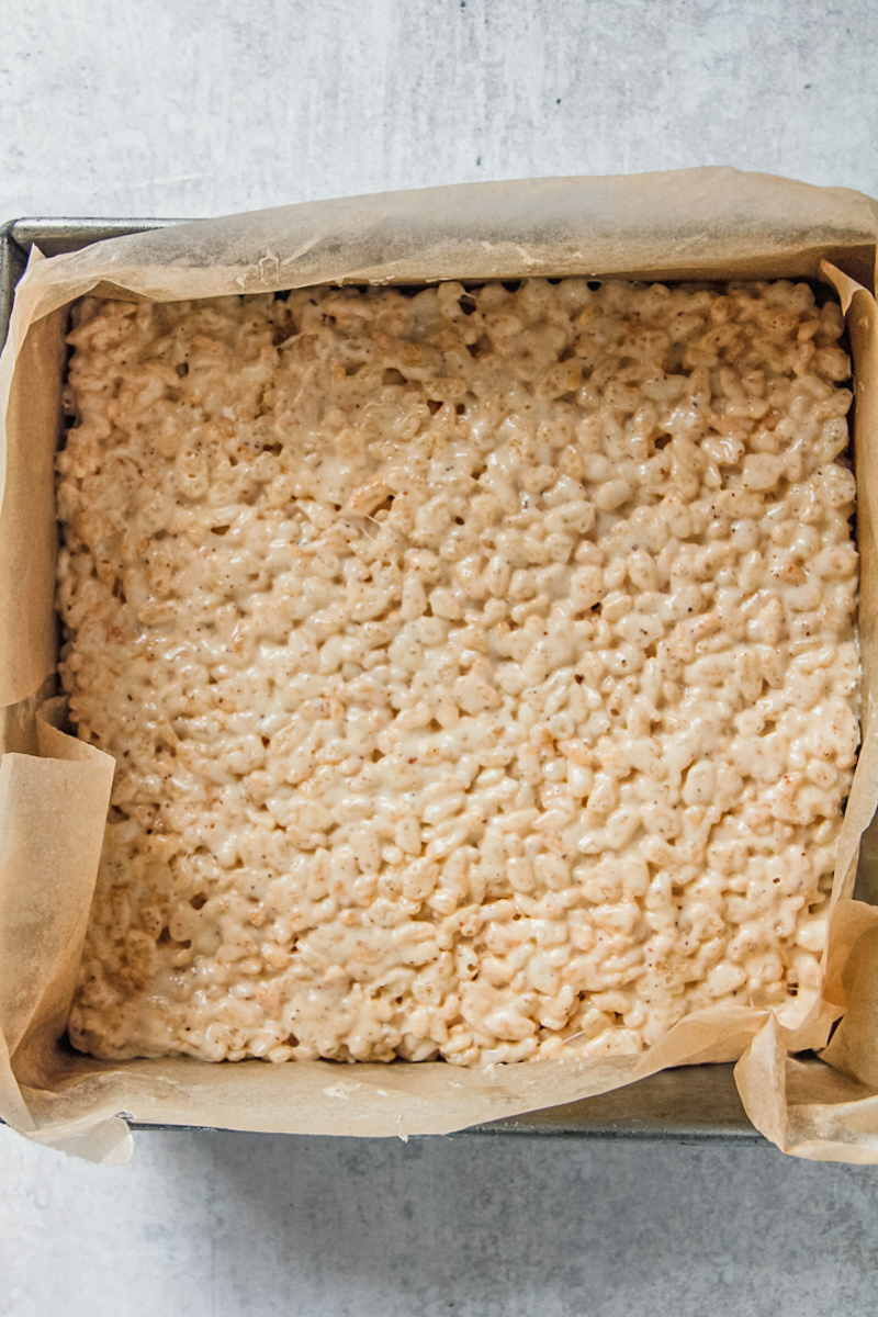 Rice krispie mixture sits pressed into a lined square metal tin on a gray surface.