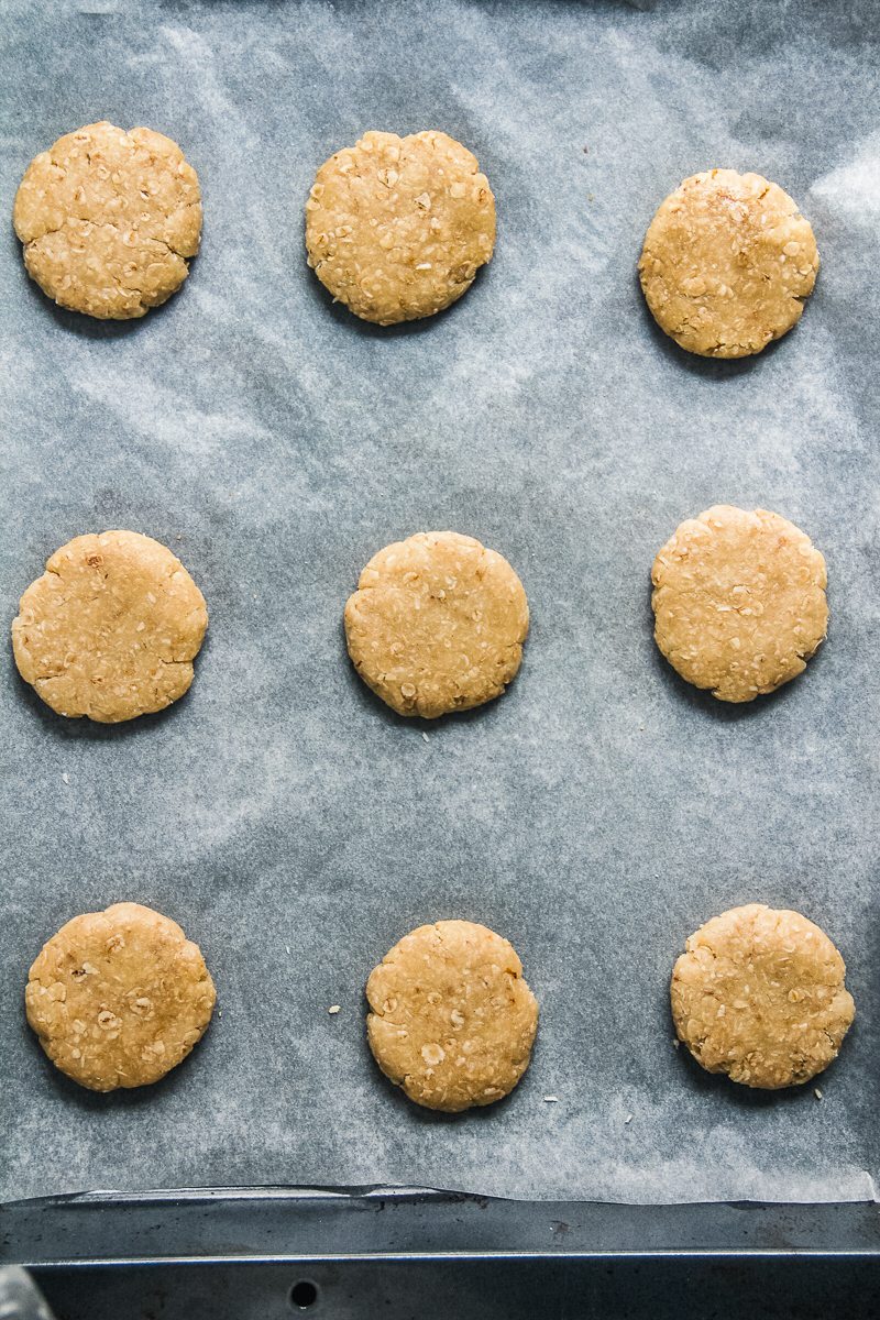 Unbaked Anzac biscuits sit on a parchment lined baking tray.